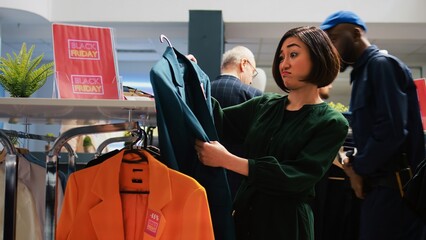 Woman customer looks through clothes hanging on racks, searching for black friday special offers in...