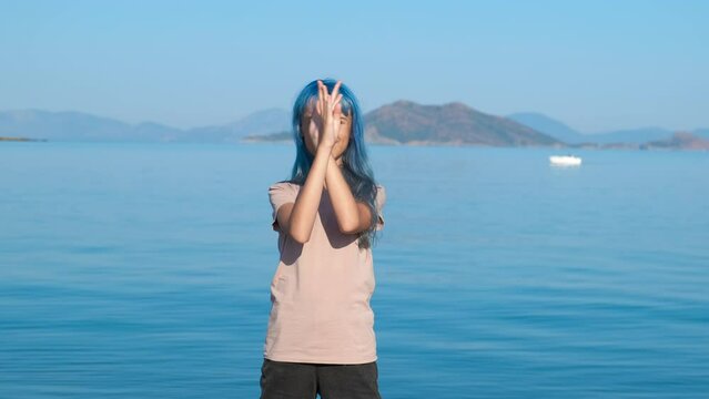 Styled teen waving hands by sea. A view of blue styled young girl dancing on the beach against sunny bay during summer vacation.