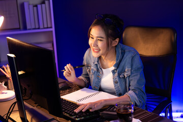 Smiling young beautiful asian creative looking on pc of profit of sales target report and writing...
