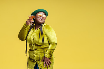 Portrait of happy stylish African American woman holding glass with  cocktail looking away isolated on yellow background, copy space. Smiling female drinking alcohol. Birthday party concept