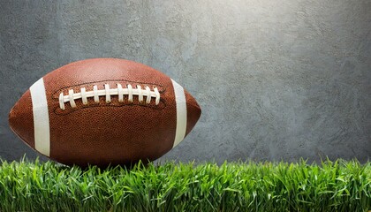 An american football ball standing over green grass in front of a gray wall. 