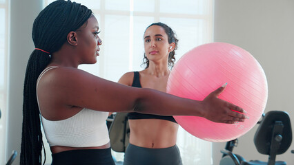 Two young woman in sportswear workout routines using exercise ball in fitness gym with trainer,...