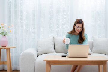 Young asian woman wearing glasses sitting sofa working on laptop computer and drinking coffee in living room at home, woman work from home with telework, freelance working, business and digital life. - obrazy, fototapety, plakaty