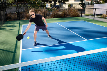 A man wearing sunglasses plays pickleball with a large shadow on a blue court