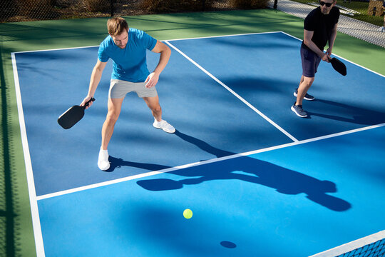 Two men play pickleball on a blue court with shadows