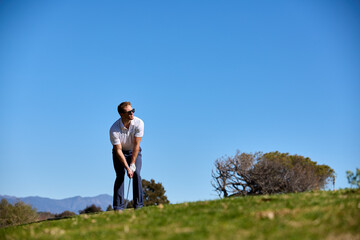 A man plays a golf shot on a golf course with blue sky behind him