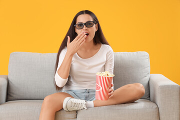 Obraz premium Surprised young woman in 3D glasses with popcorn watching movie on sofa against yellow background