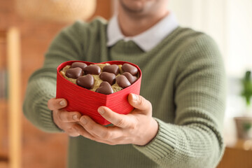 Young man with box of heart-shaped chocolate candies at home, closeup