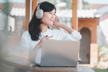 A contented young Asian woman with headphones and a smartphone enjoying music while working on her laptop outside.