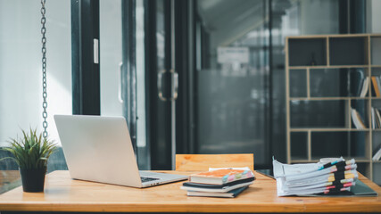 A tidy office desk displaying an open laptop, a small potted plant, and a stack of documents with colorful tabs in a well-lit workspace with bookshelves. © Satori Studio