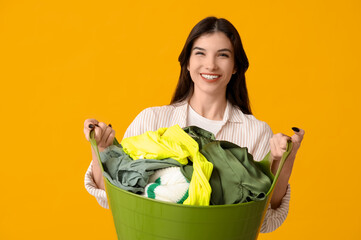 Happy young woman holding basket with clean laundry on yellow background