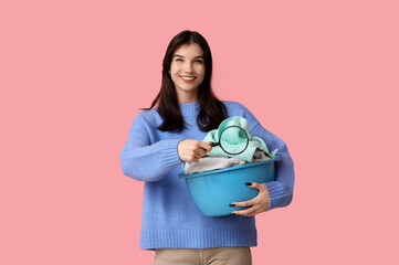 Beautiful young woman holding basket with clean laundry and magnifier on pink background