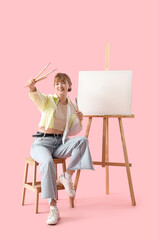 Female artist with paint brushes and easel on pink background