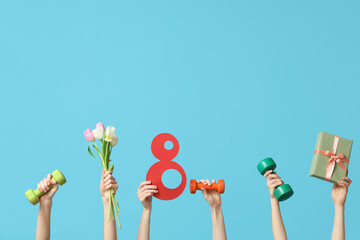 Female hands with paper figure 8, dumbbells, gift box and flowers for International Women's Day on color background