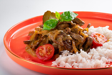 Beef in cream sauce with onions in a red plate with rice on a white background. A dish of beef...