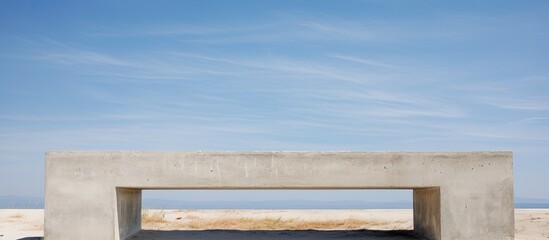 A concrete bench is positioned on top of a sandy beach under a clear blue sky. The sturdy bench contrasts with the soft sand, offering a place to sit and enjoy the coastal scenery.