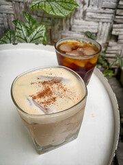 A glass of iced coffee and caffe latte drinks on a wooden table with houseplants background. Black coffee with ice cubes on the surface of the drink. Americano Long Black and Milk Coffe.