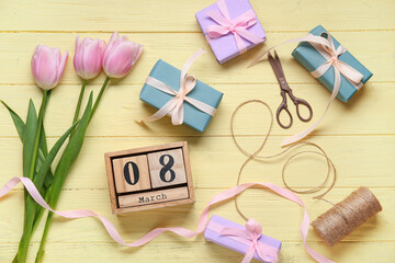 Cube calendar with date MARCH 8, gift boxes, pink tulips and scissors on yellow wooden background. International Women's Day celebration. Top view
