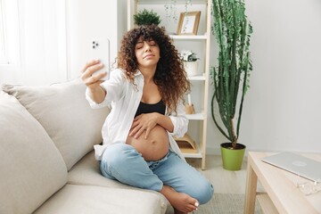 Pregnant woman blogger sits on the couch at home and takes pictures of herself on the phone, selfie and video call, consultation with the doctor online, pregnancy management