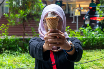 An Asian Muslim woman holding an ice cream cone with both hands. Outdoors.