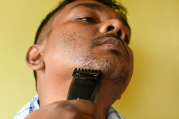 A closeup of a young Indian man trimming his beard with an electric trimmer. Selective focus on...