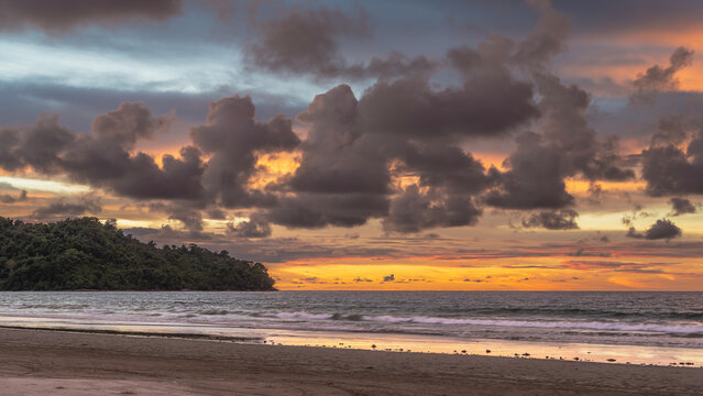 A bright tropical sunset. Purple clouds float in the orange-lit sky. The waves of the turquoise ocean foam on the sandy beach. A green hill in the distance. Madagascar. Nosy Be 