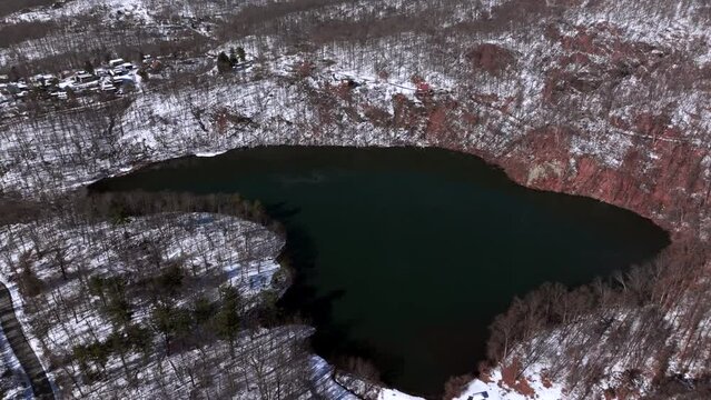 An aerial view of a large emerald colored lake, surrounded by mountains during winter on a sunny day. The camera boom up then truck right, tilted downwards orbiting the lake.
