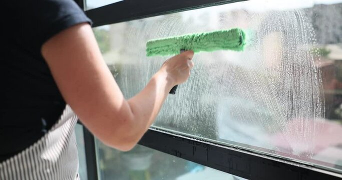 Woman in apron carefully cleans a window with scraper and sprayer