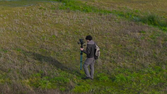 Male Photographer Carrying Camera On Tripod On The Field Near Lake. aerial shot