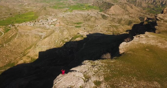 A man dangerously sitting on the edge of a cliff abyss rock headland with the view of rural village countryside in Dezful Iran natural landscape of highland riverside a nature adventure hiking aerial