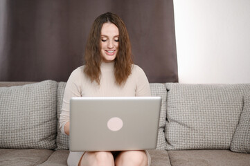Woman working on laptop in home office while sitting on sofa. Young female student studying...