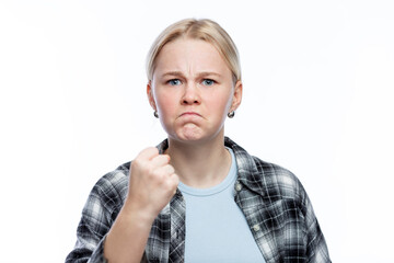 Frowning teenage girl shows her fist. A cute blonde with freckles on her face wearing a plaid shirt and a blue top. Negative emotions and aggression. White background. Close-up.