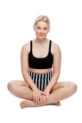 A smiling teenage girl in a sports swimsuit sits on the floor. A cute blonde with freckles on her face in a black top and striped high pants. Isolated on a white background. Vertical.