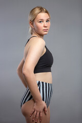 Beautiful teenage girl in a sports swimsuit. A cute blonde with freckles on her face in a black top and striped high pants. Gray background. Vertical.