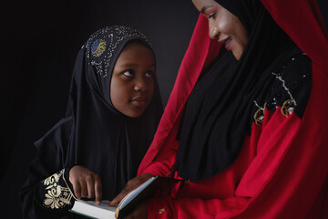 African Muslim mother in traditional costume teaching her daughter to read the Quran, little girl...