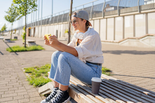Young Hispanic hipster girl browsing mobile phone while sitting in street with takeaway beverage. 