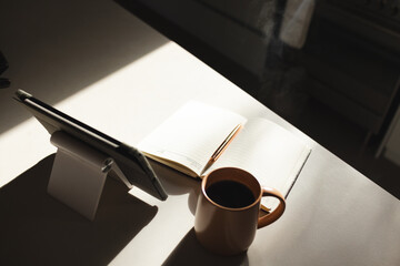 A tablet with a stylus, an open notebook, and a cup of coffee rest on a sunlit table with copy space