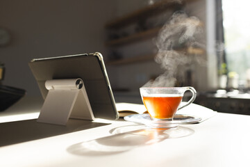 Steam rises from a hot cup of tea next to a tablet on a sunlit table