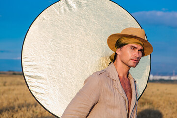 Portrait of a tall handsome man dressed in a coarse linen suit and hat standing at golden oat field with a golden reflector on the background.