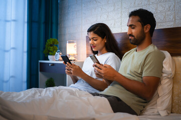 Relaxed Indian couples busy using mobile phones on bedroom before sleeping at night - concept of...