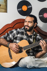 Latin man playing acoustic guitar in his room. Musician singing while using harmonic instrument