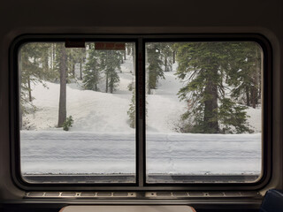 Fototapeta premium Train window view of snowbanks rolling by in a hilly, snowy forest. The interior shows booth seating, with seats on either side of a table for passengers to eat dinner. The Sierra Nevadas are outside.
