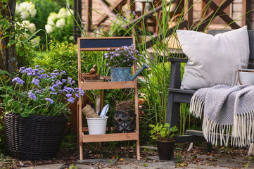 Summer outdoor backyard decorations. Rustic flower stand with potted plants, wooden bench with pillows and blanket. Cozy garden relax area.