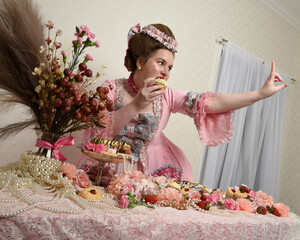 Close up portrait of cute female model wearing an opulent pink gown, the costume of a historical French baroque nobility. Eating indulgent food feast with pastries, flowers and rich jewels