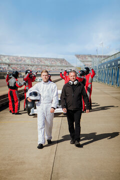 Two men confidently walk down a racetrack with a team of pit crew members in the background. 
