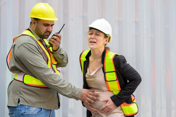 Male engineer in yellow helmet using walkie talkie to request emergency vehicle for pregnant manager. Caucasian woman in white helmet suffering belly ache while working with colleague  at cargo yard - 747028814