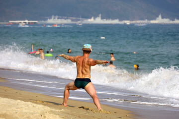 Yoga on the beach, muscular man stands on sea waves background. Balance with nature, fitness and exercise at vacation