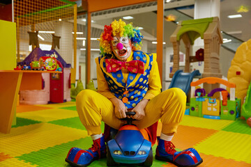 Overjoyed clown having fun riding toy car at playroom