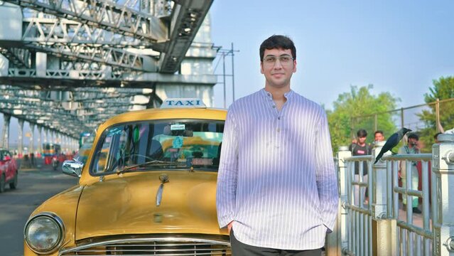 A young confident handsome Indian Asian man or male wearing eyeglasses and standing in front of an iconic and resting on a vintage yellow cab looking at the camera and smiling in Kolkata city
