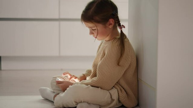 Close-up of a little brown-haired girl in a cream sweater sitting on the floor near the kitchen table and doing something on the phone during her time alone in the evening